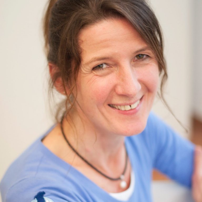 Frau mit lächelndem Gesicht, braunen Haaren und blauer Bluse, sitzt in einem hellen Raum.